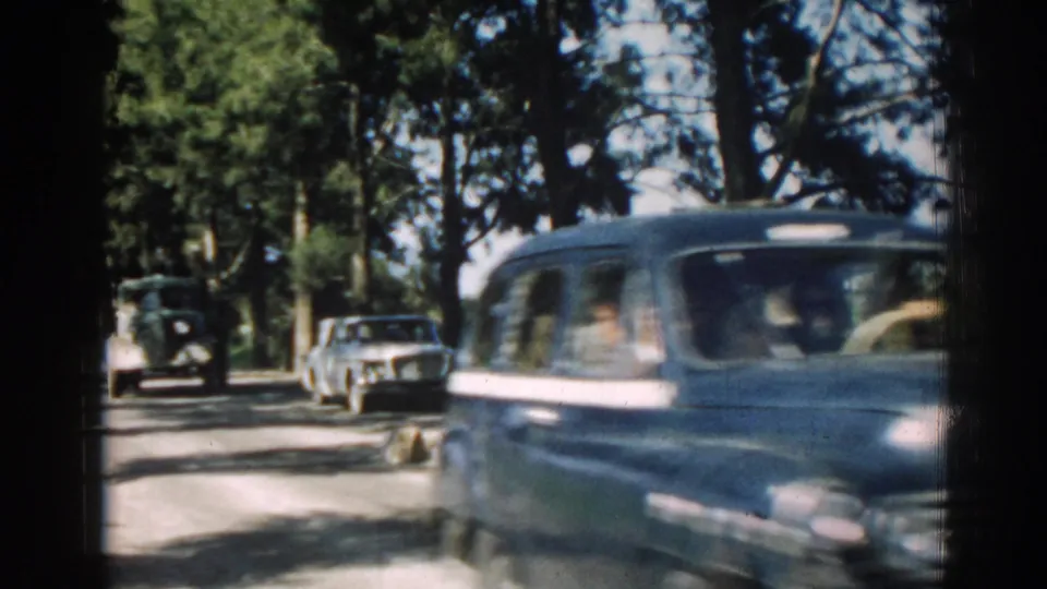 A blue mid-century car passes along a shaded Izmir road, with other vintage vehicles visible farther back.
