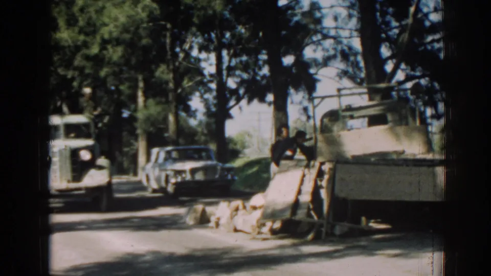 View past the truck bed toward passing cars on a shaded road in 1962 Izmir, with rocks piled near the shoulder.