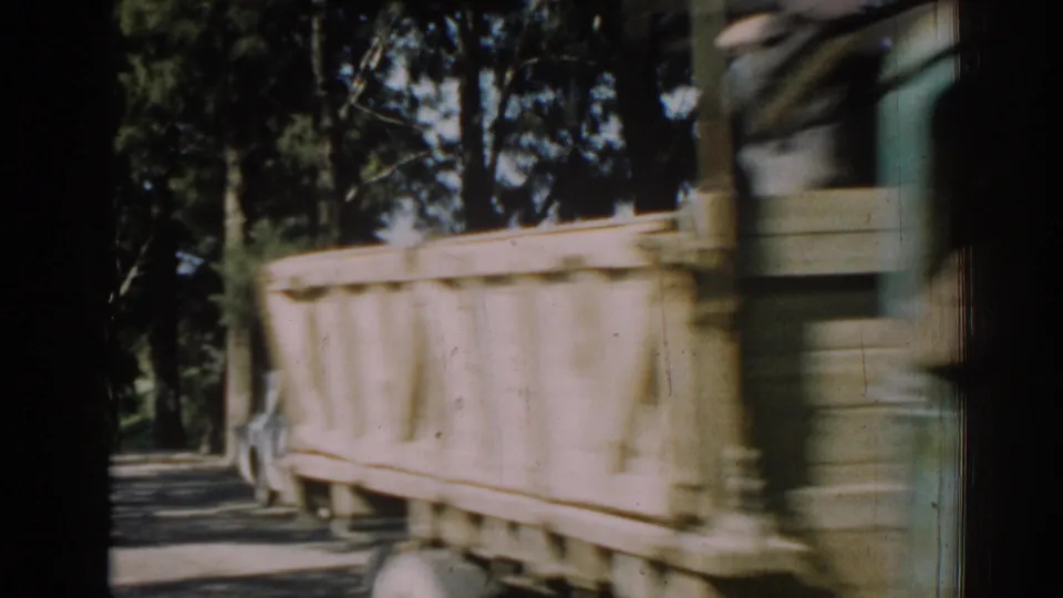 A green open-bed truck passes close to the camera on a sunny Izmir road, with motion blur and trees behind it.
