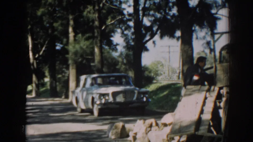 A white sedan approaches or passes on a shaded Izmir road while a person stands near a wooden roadside structure.