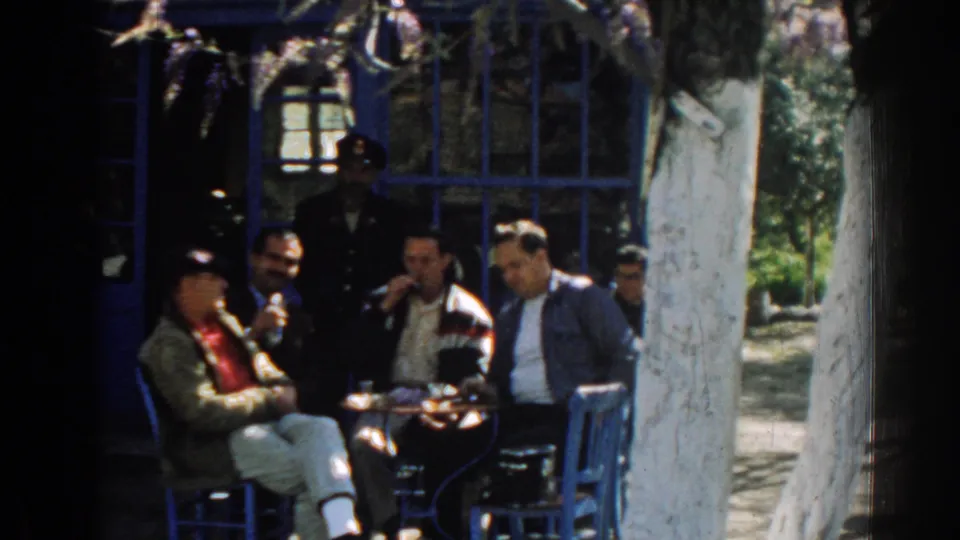 In 1962 Izmir, Turkey, several men sit and stand around a small table outside a blue-painted building with window frames and chairs.