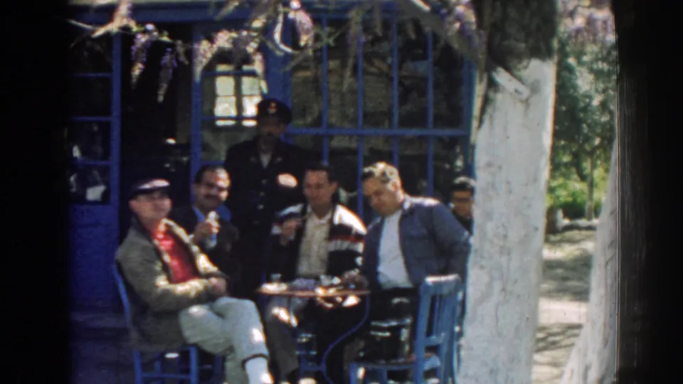 A group of men in 1962 Izmir sit outdoors with glasses on a small table, framed by blue-painted window trim and dappled shade.