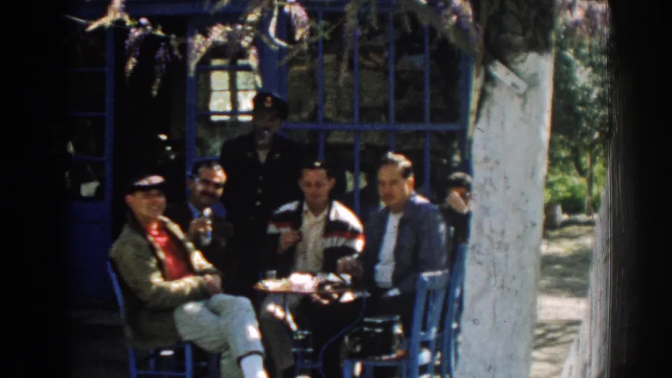The 1962 Izmir scene continues with men gathered around a small outdoor table beside blue-painted windows and a pale tree trunk.