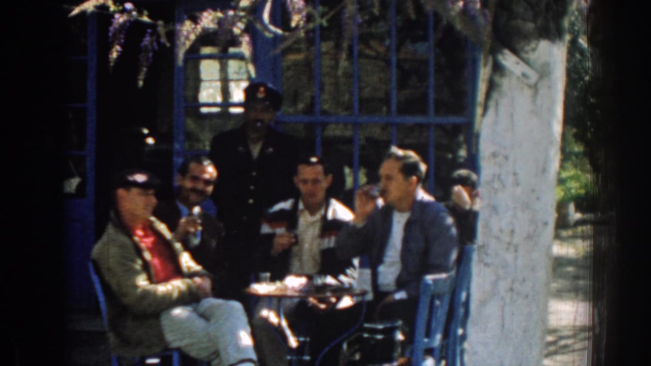 Four men sit around a small outdoor table while a uniformed man stands behind them.