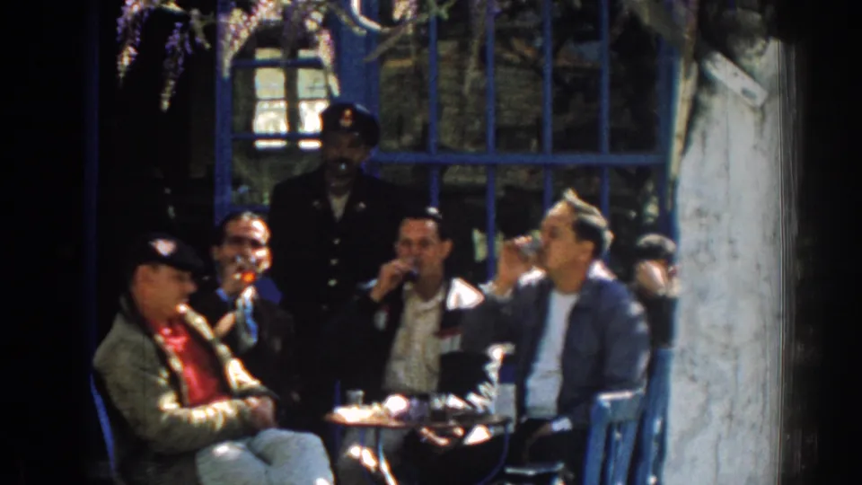 A 1962 Izmir home-movie frame shows men at an outdoor table drinking from small glasses, with blue window bars and a uniformed man behind them.