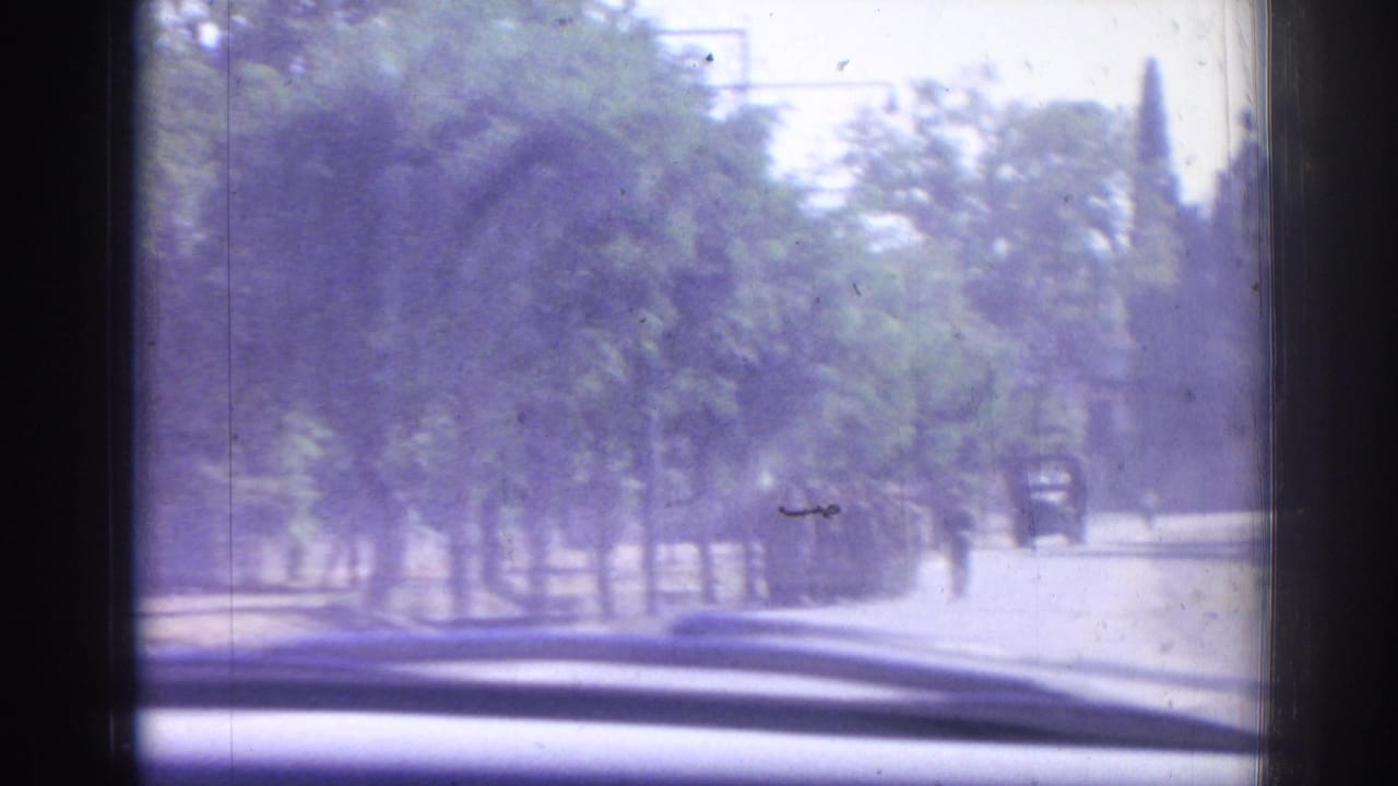 View through a car windshield toward a tree-lined road with distant soldiers and a vehicle.
