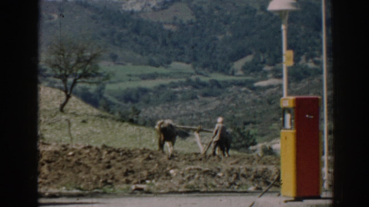 Farmer and oxen plowing a hillside field beside a red and yellow roadside pump.