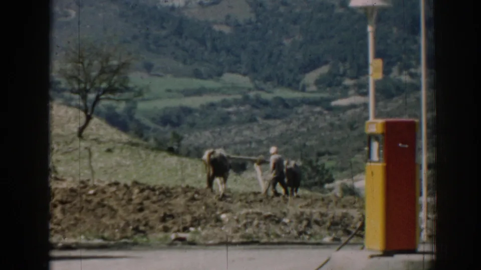 1962 Izmir frame with a farmer and two oxen moving across tilled ground, with hills and a pump nearby.