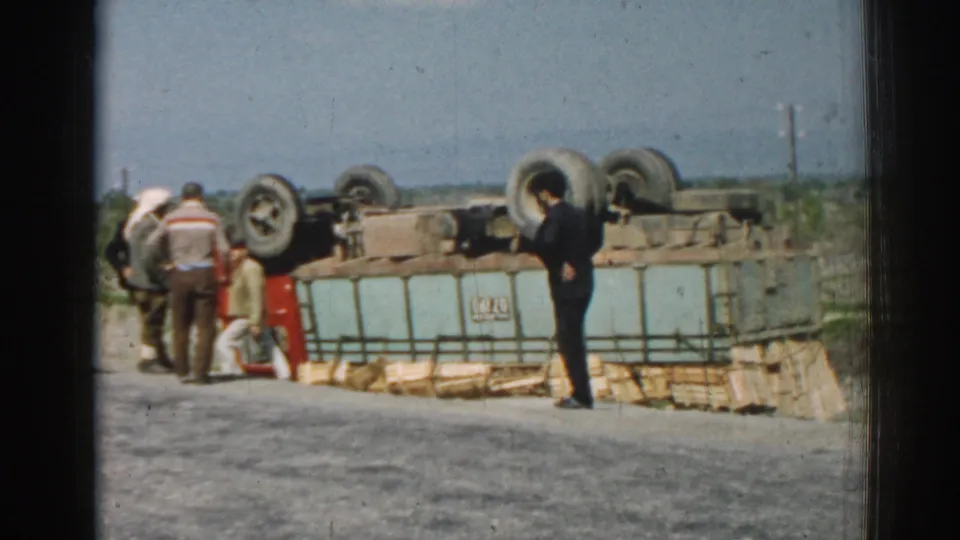 1962 Izmir roadside frame with men gathered around an overturned red and light-blue truck and scattered cargo along the road edge.