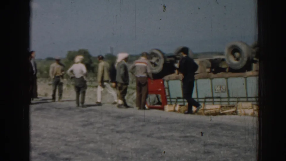 1962 Izmir home-movie frame showing several men gathered near an overturned truck, with rural land and sky beyond the road.
