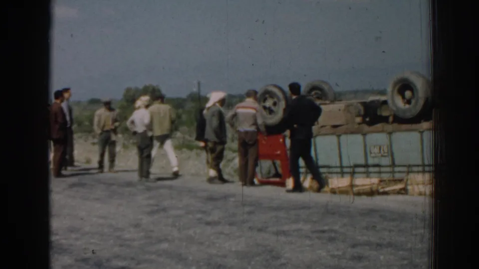 In 1962 Izmir, Turkey, several adult men gather near an overturned red truck on a rural roadside.