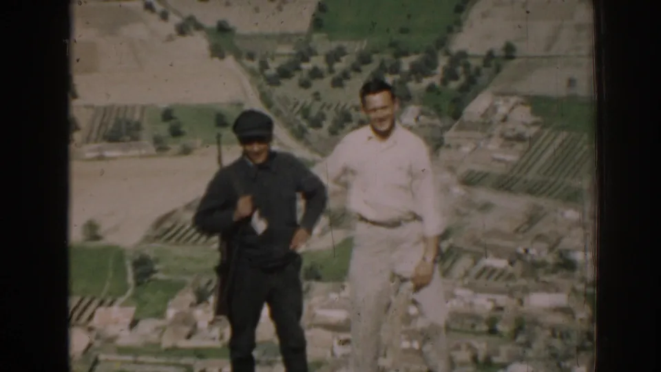 Two adult men, one in dark clothing and one in light clothing, stand in a rural Izmir landscape with fields and trees below.