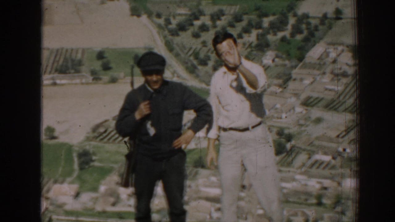 Civilian in light clothing gestures while a uniformed man with a rifle stands beside him.
