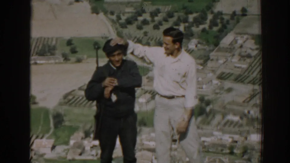 On a hilltop in Izmir, the civilian reaches toward the cap of the uniformed man holding a rifle, with fields behind them.