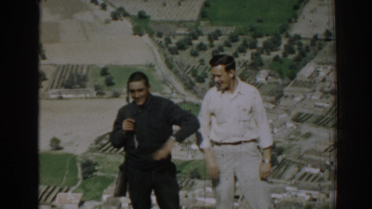 Two men standing on a hillside with fields and buildings below.