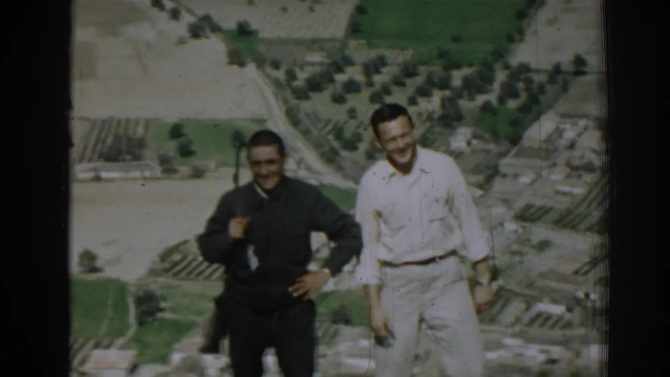 Two men smile toward the camera on a high overlook near Izmir, with cultivated fields, groves, and roads visible below.