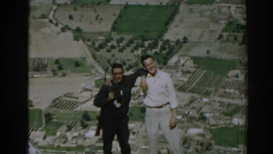Two men laugh and lean close on a hillside in 1962 Izmir, with a patchwork agricultural landscape stretching behind them.