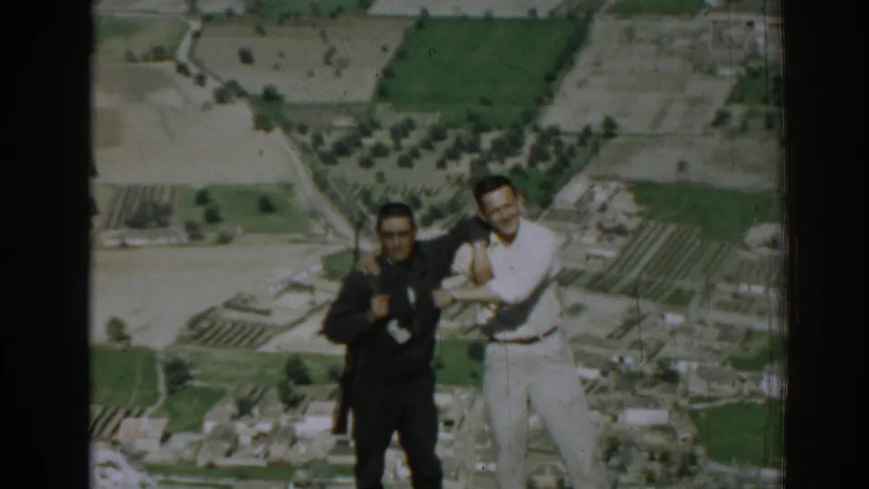 Two men pose closely on a hilltop near Izmir, Turkey, with farmland, groves, roads, and small buildings below.