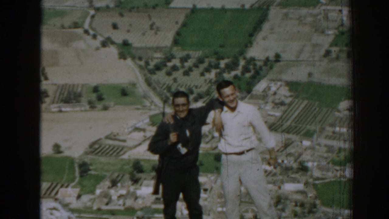 Two adult men smile and stand together above Izmir, Turkey, with fields, groves, and buildings below in 1962 home-movie footage.