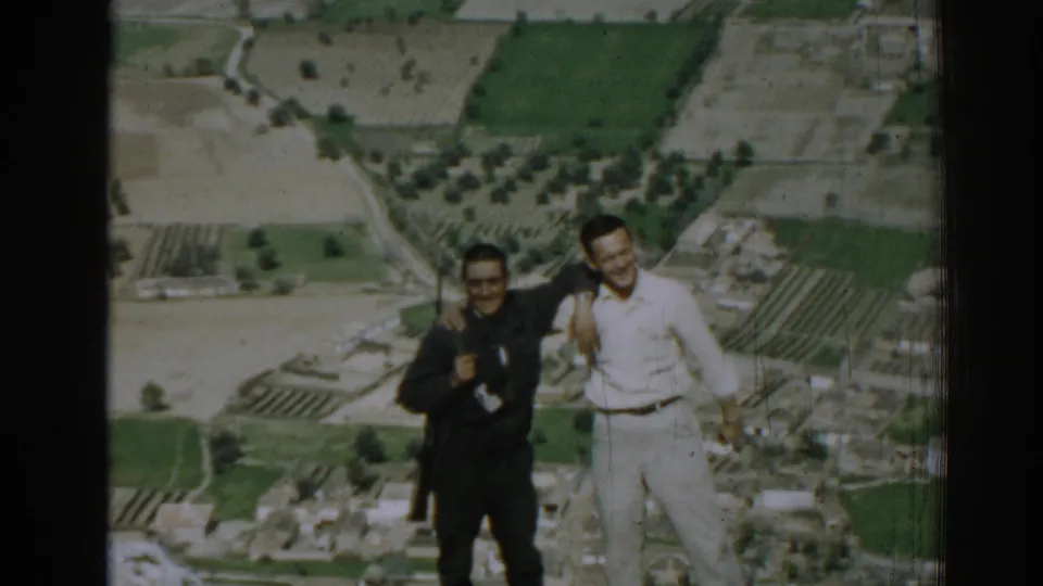 Two adult men smile and stand together above Izmir, Turkey, with fields, groves, and buildings below in 1962 home-movie footage.
