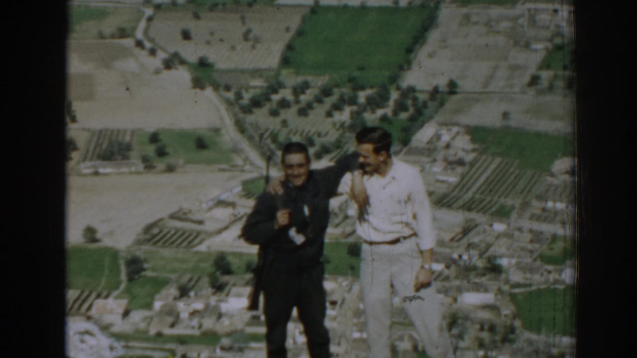 Two adult men stand arm in arm against a distant landscape of fields and buildings.