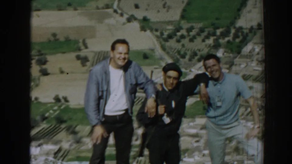 Three adult men smile and lean together at an elevated Izmir viewpoint, with 1960s fields and urban edges in the background.