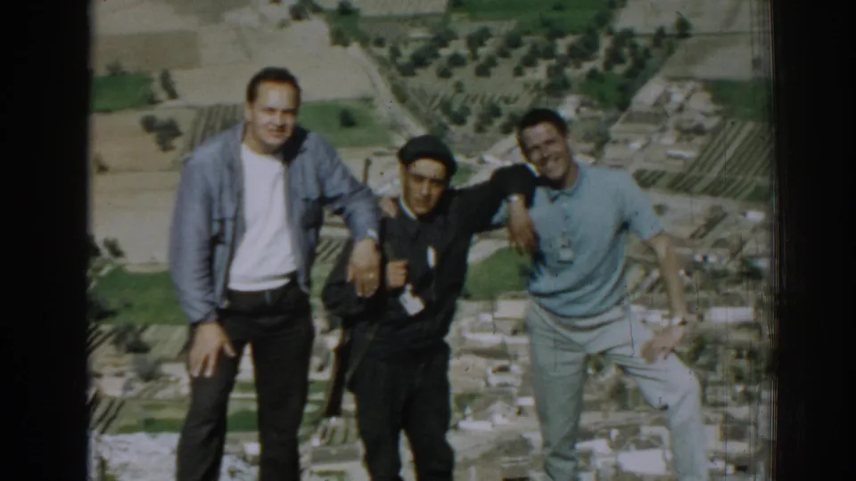 Three adult men pose casually for a 1962 home movie above Izmir, with one holding a camera and fields visible below.