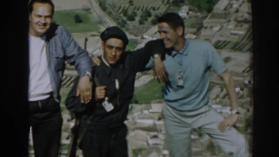 Three young men in casual clothing, one wearing a dark beret, stand close together above rural Izmir farmland.