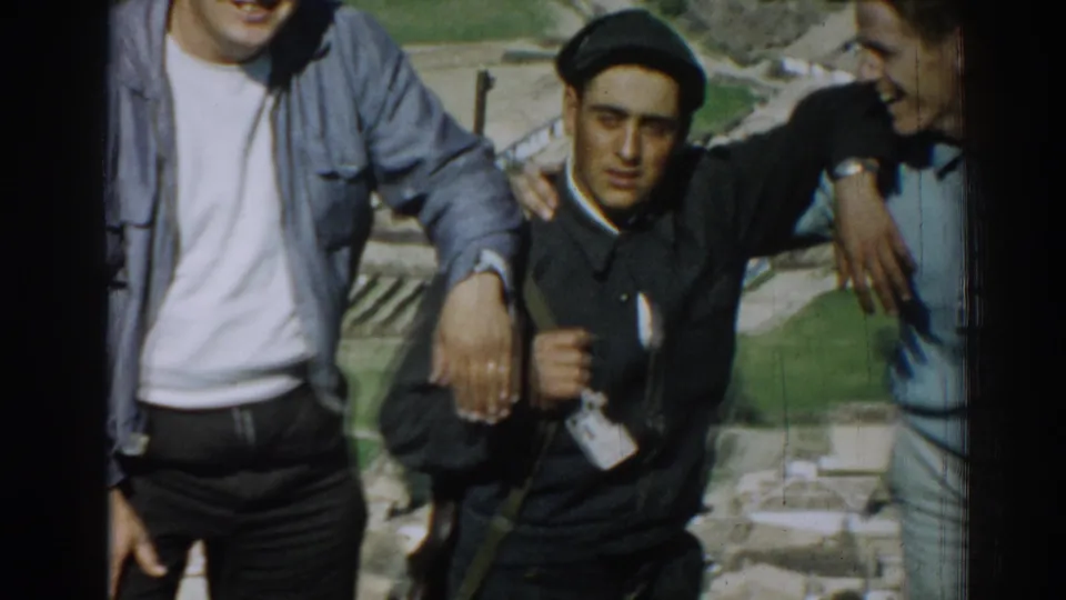 A close home-movie frame centers the young man in a dark beret between companions above Izmir farmland.