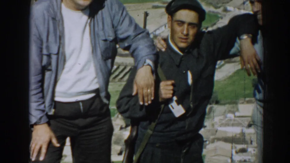 In 1962 Izmir, Turkey, three adult men pose on a sunny elevated site, with the central man in dark clothing and cap partly framed by two companions.