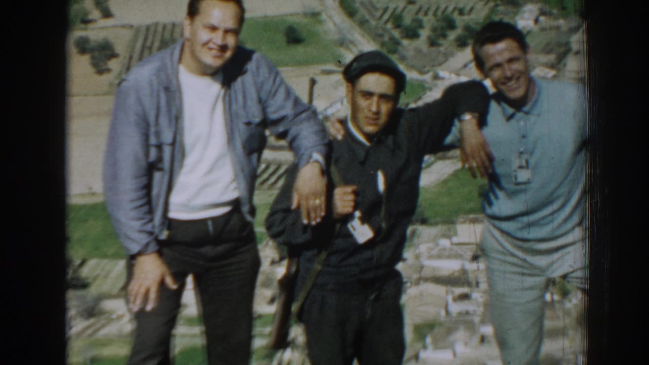 Three men stand arm in arm on a hillside, the central man wearing a dark cap and holding a rifle-like object.