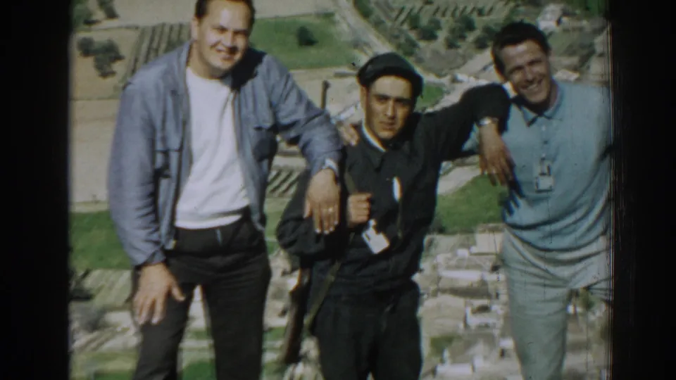 Three adult men pose together in bright sun in Izmir, Turkey, with the central figure in dark clothing and two casually dressed companions.