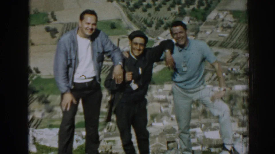A 1962 home-movie frame shows three men smiling on an elevated outdoor site near Izmir, with the central man in dark clothing and cap.