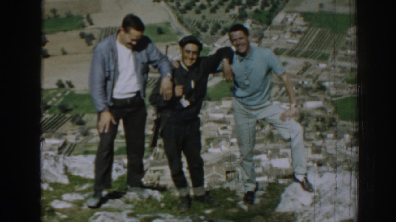 Three adult men pose on rocky ground above a valley with ruins nearby.