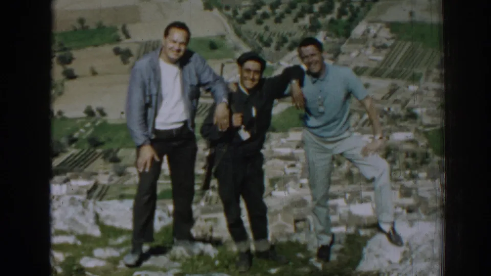 Three men in casual early-1960s clothing smile at the camera on a high Izmir overlook with ruins and fields behind them.