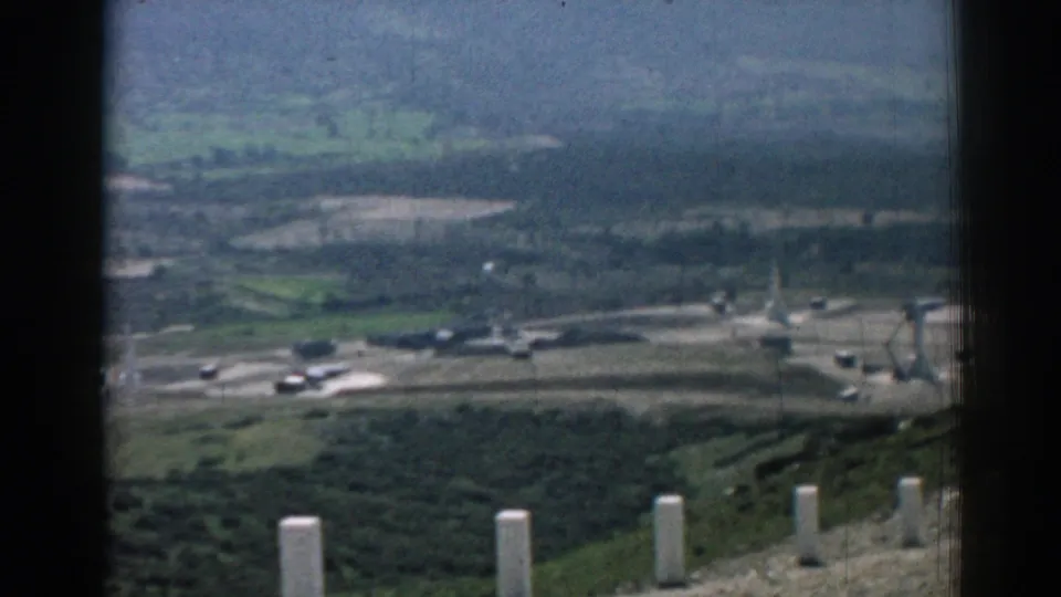 From an elevated Izmir overlook, the frame shows a distant roadway, low buildings, vehicles, and rolling green terrain.
