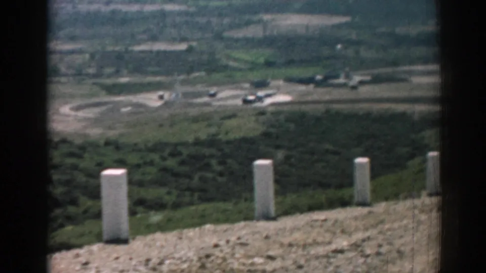 A 1962 home-movie frame from Izmir shows rocky roadside ground, white posts, shrubs, and a distant built area in the valley.