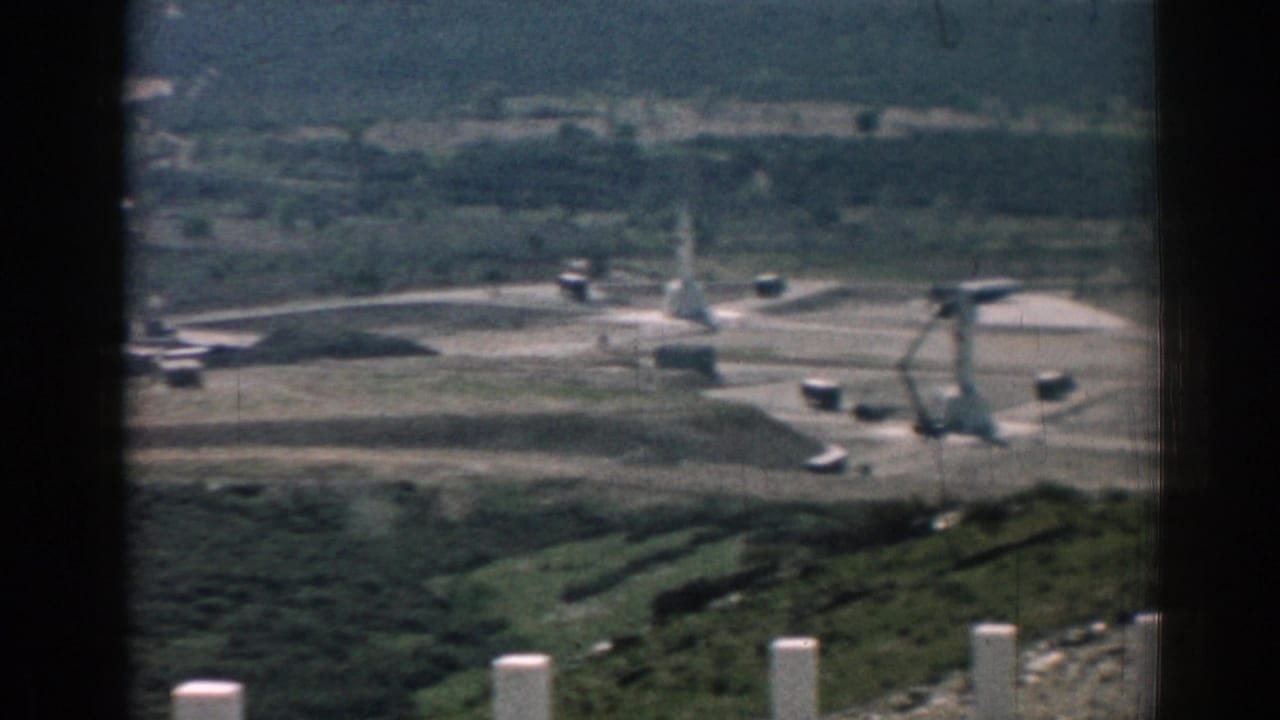Distant hillside construction site seen past dark film gate edges and foreground posts.