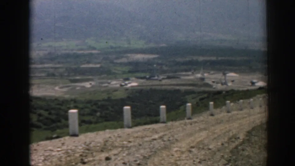 1962 Izmir home-movie view from a ridge road, with white concrete posts, dry hillside, green valley fields, and distant structures.