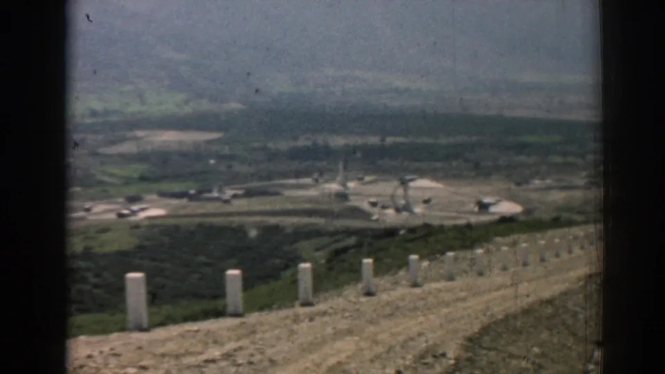 A soft 8mm frame shows the ridge road edge above Izmir's valley, with posts, hillside scrub, fields, and distant industrial-looking structures.