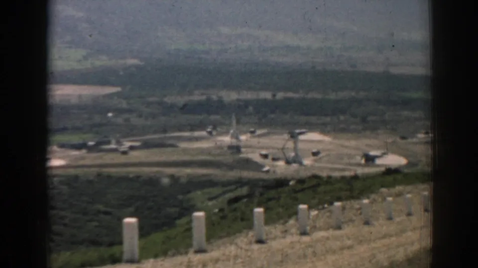 Late in the clip, the 1962 Izmir ridge road view looks past white posts toward a valley with fields, scattered structures, and muted hills.