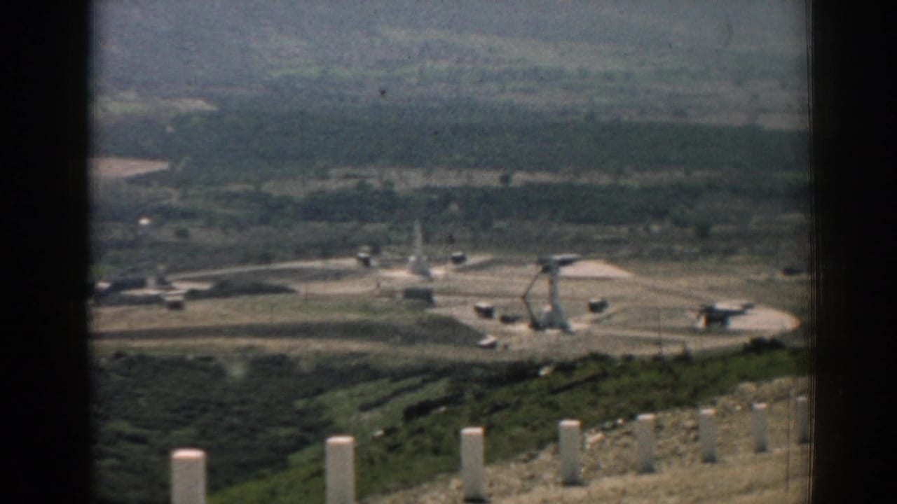 Distant missile site seen through dark window-frame edges with launcher equipment and hills beyond.