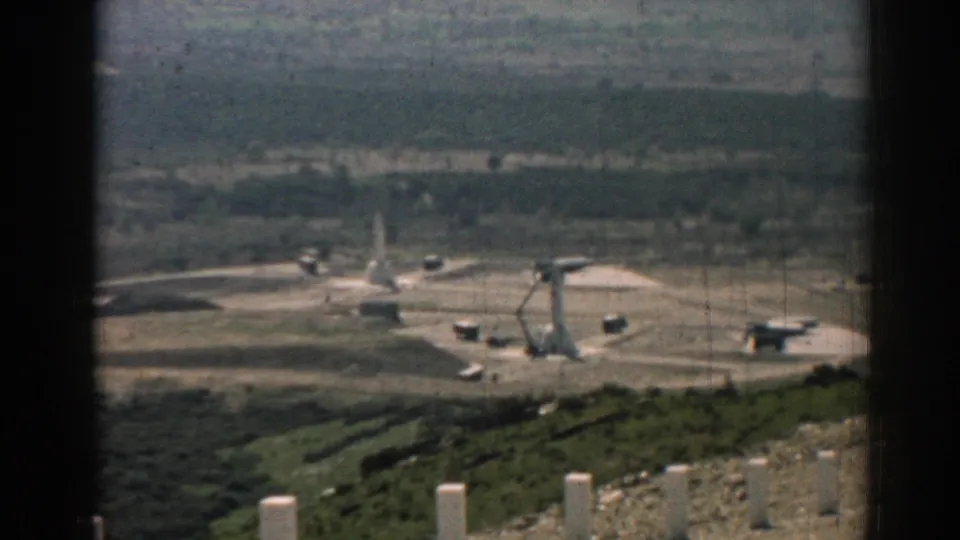 A distant 1962 Izmir view shows launcher equipment and vehicles on a pale pad, with hills and dark frame edges around the shot.