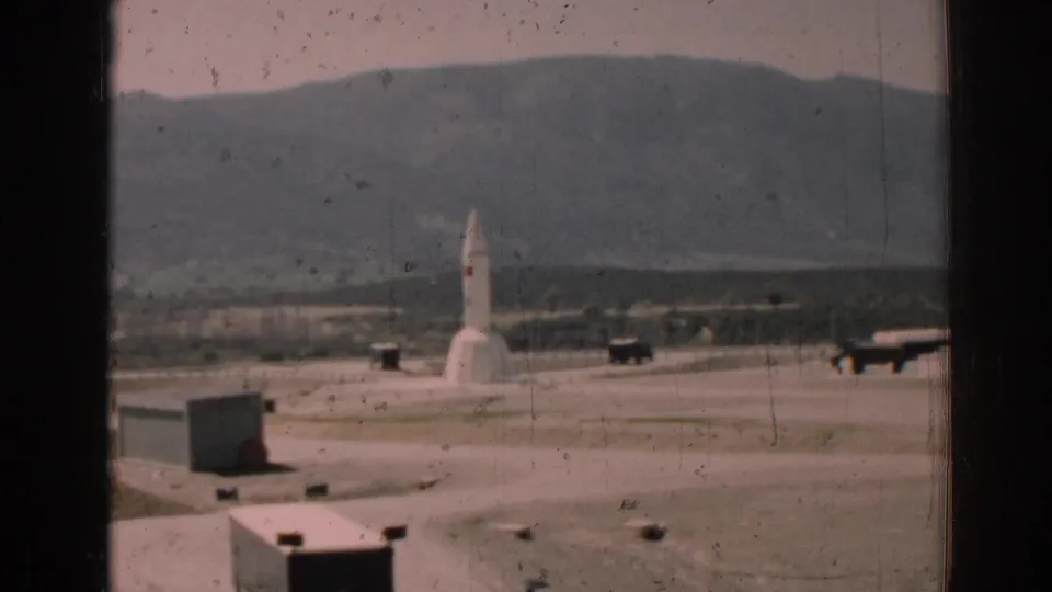 White missile at a 1962 Izmir launch site, filmed from a distance with support vehicles, concrete ground, and hazy hills behind it.