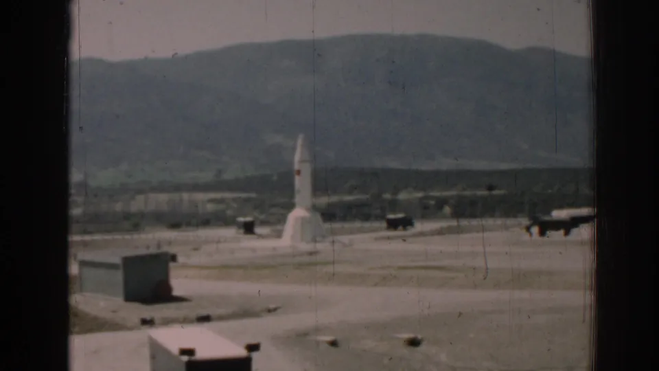 1962 Izmir, Turkey footage showing a white rocket-like structure on a paved compound, seen through dark window framing with hills beyond.