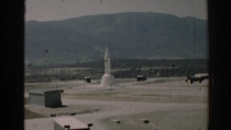 The rocket remains centered at the outdoor facility, with small vehicles or equipment and a hilly landscape visible behind it.