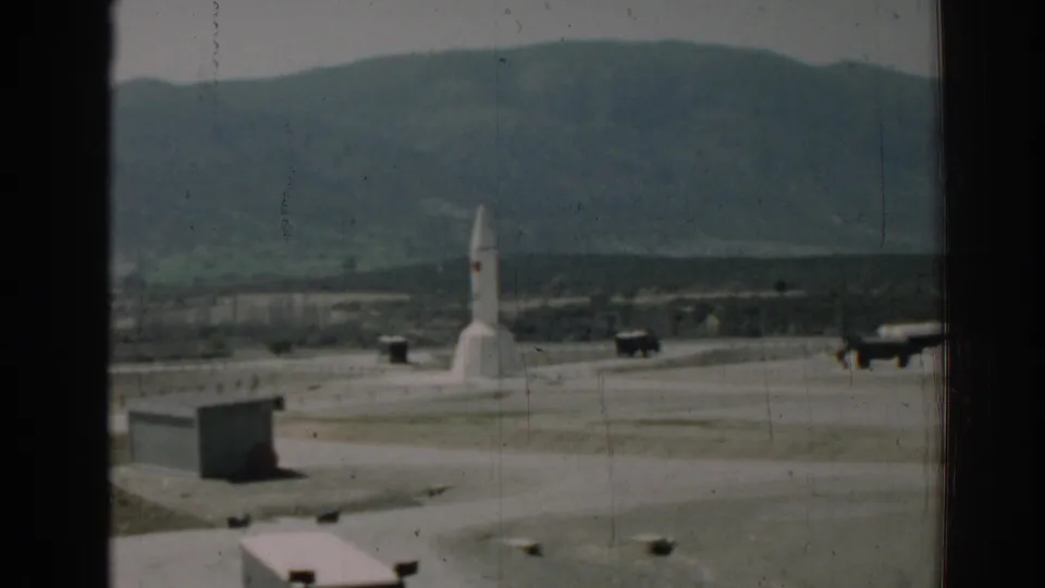A softer 8mm view shows the white rocket on the paved facility grounds, partially divided by dark foreground framing.