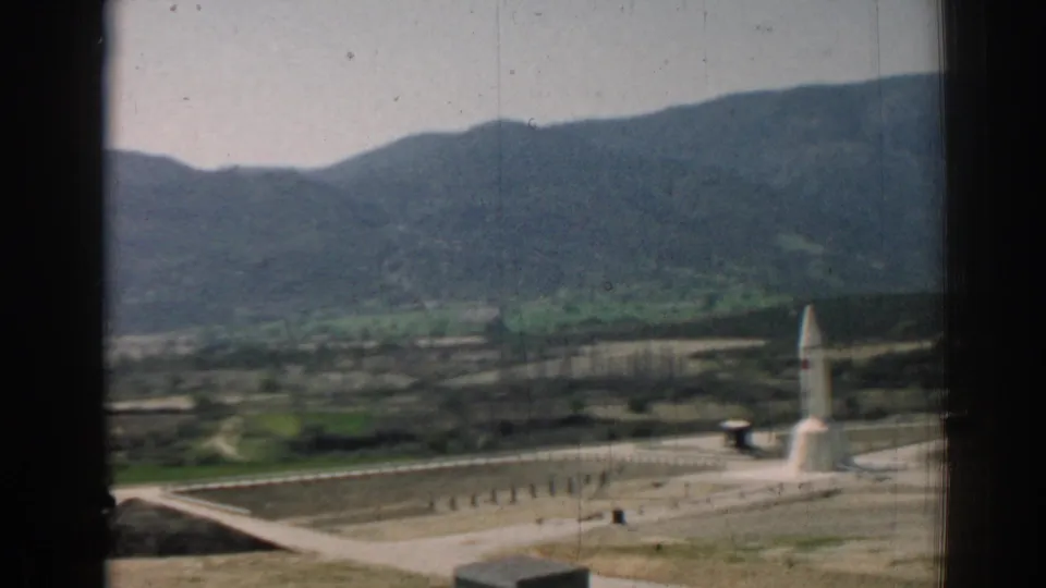 Distant 1962 8mm view near Izmir showing a white rocket-like object on a paved site, partially framed by dark window edges.