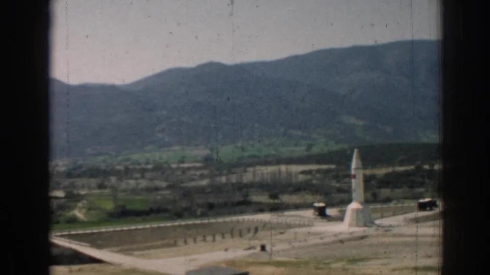 Faded 1962 8mm frame from Izmir, Turkey, showing a rocket-like structure on open grounds below dark green mountains.
