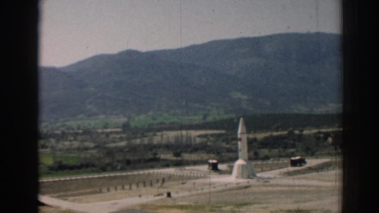 Window-framed view of a white monument on sandy ground with hills and mountains behind it.